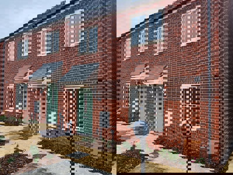 A two-storey red brick housing development with a sage-coloured front door and newly planted flower beds