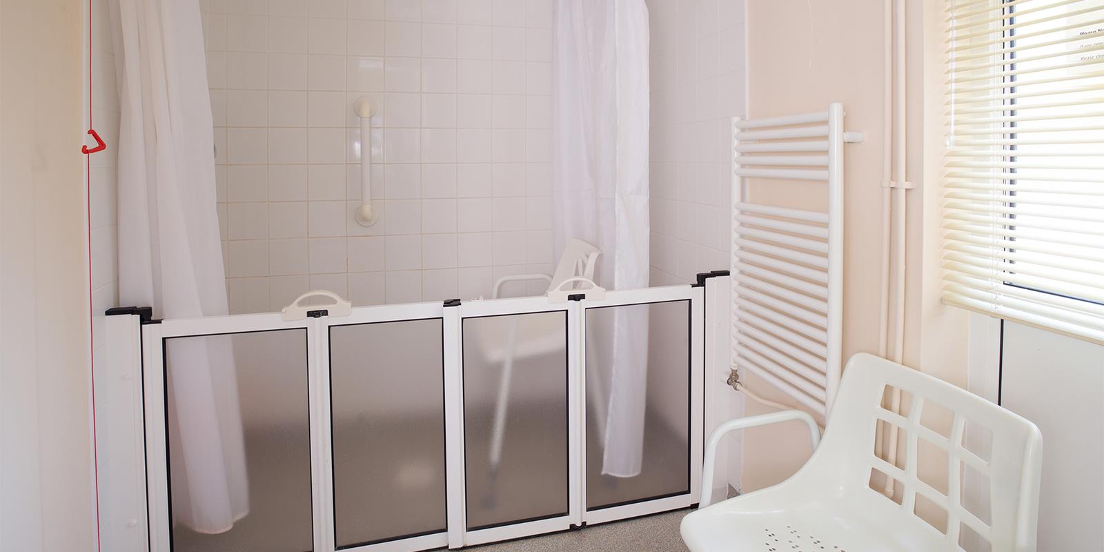 A shower-room with a walk-in shower, featuring a retractable plastic gate and a white plastic chair in the foreground